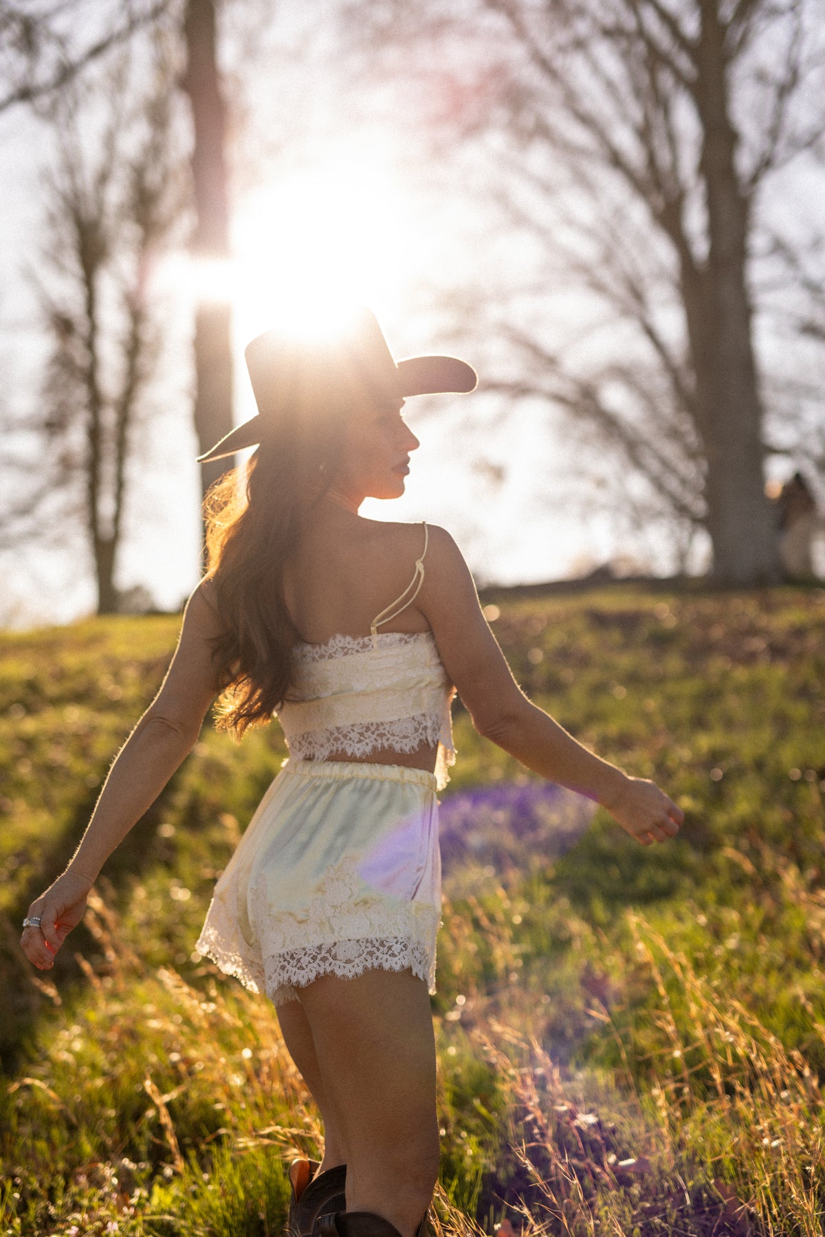 Woman in a lace outfit and cowboy hat standing in a field with the sun shining through.