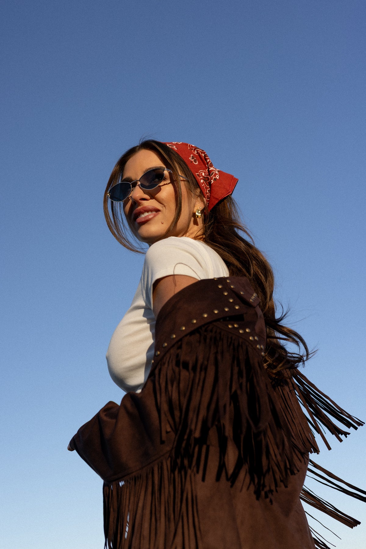 Woman wearing a brown fringed jacket and red bandana against a clear blue sky