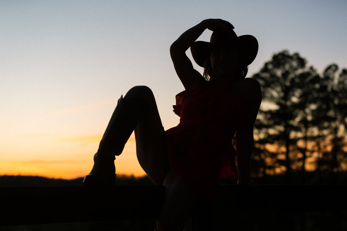 Silhouette of a woman sitting with a hat on against a sunset sky