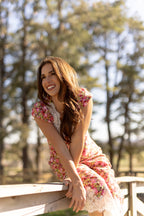 Woman in a floral dress sitting on a wooden railing with trees in the background