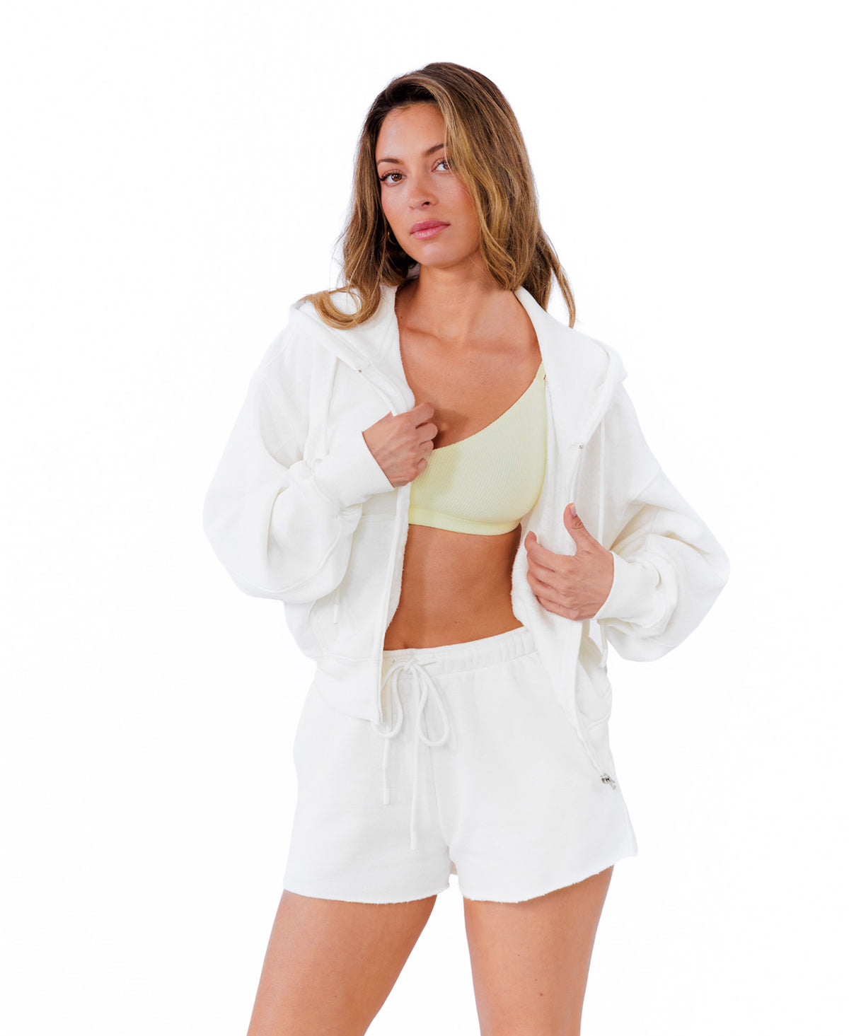 A woman modeling a white zip-up hoodie and matching white drawstring shorts over a pale yellow sports bra, photographed against a solid white background.