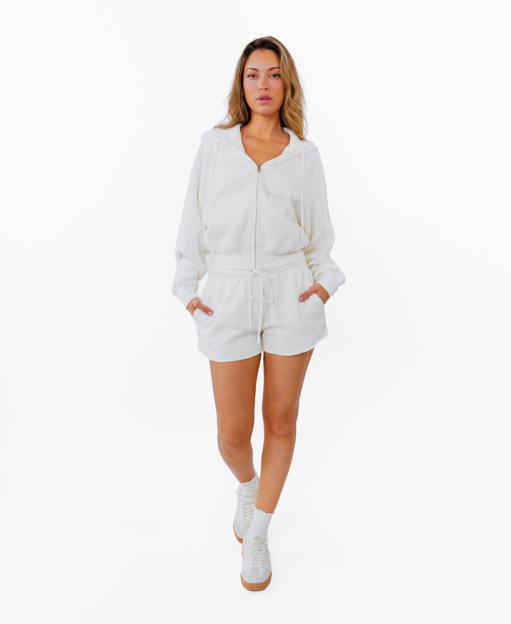 A woman modeling a white zip-up hoodie and matching white drawstring shorts, photographed against a solid white background.