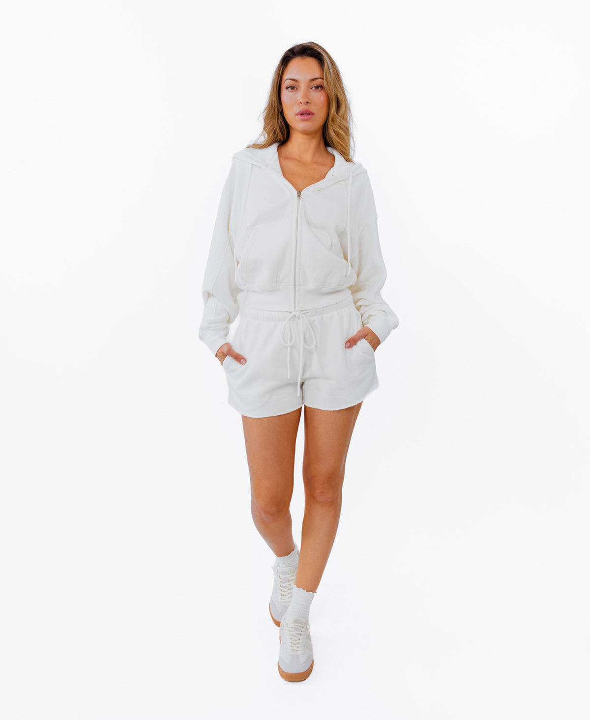 A woman modeling a white zip-up hoodie and matching white drawstring shorts, photographed against a solid white background.
