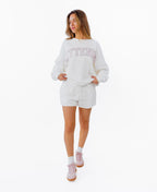 A woman modeling a white crewneck sweatshirt with 'KITTENISH' printed in pink arched letters across the chest, paired with matching white shorts, white socks, and pink sneakers, set against a plain white background.