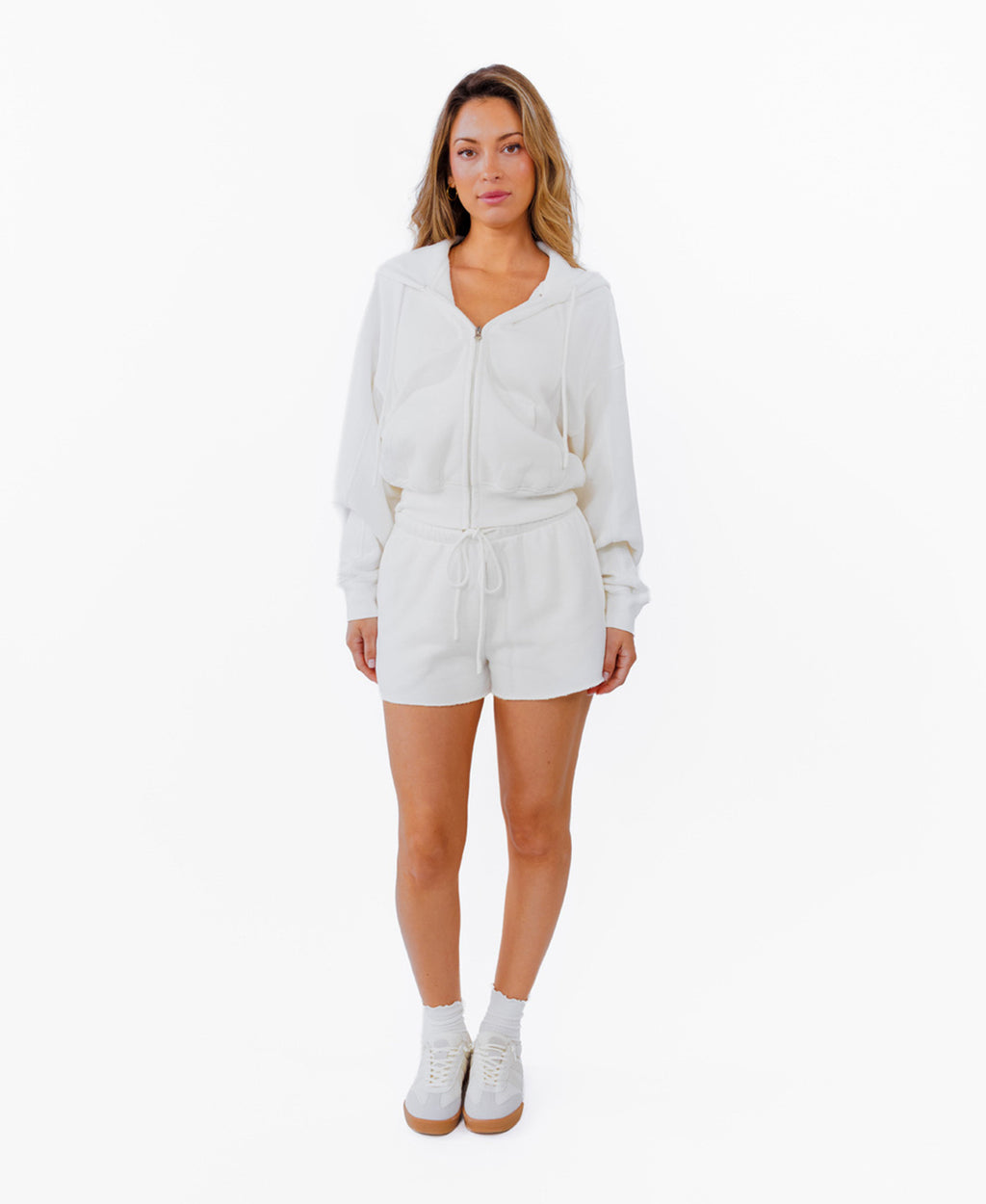A woman modeling a white zip-up hoodie, white drawstring shorts and white sneakers against a plain white background.