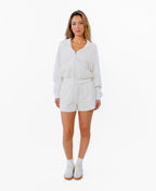 A woman modeling a white zip-up hoodie, white drawstring shorts and white sneakers against a plain white background.