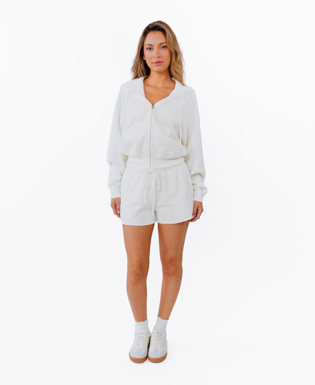 A woman modeling a white zip-up hoodie, white drawstring shorts and white sneakers against a plain white background.