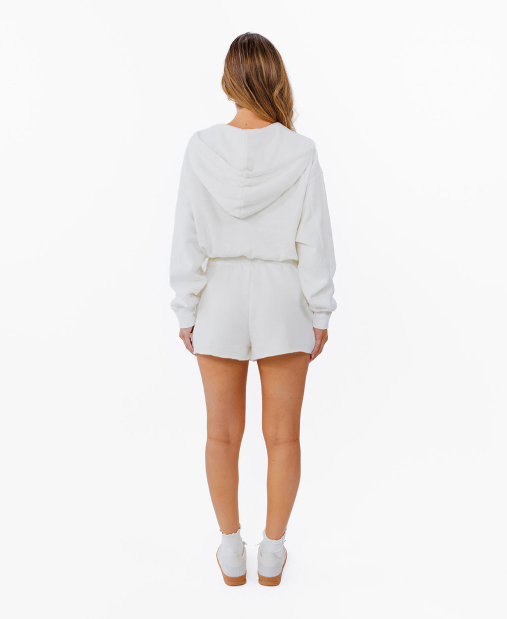 A woman modeling a white zip-up hoodie, white drawstring shorts and white sneakers against a plain white background.
