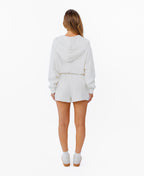 A woman modeling a white zip-up hoodie, white drawstring shorts and white sneakers against a plain white background.