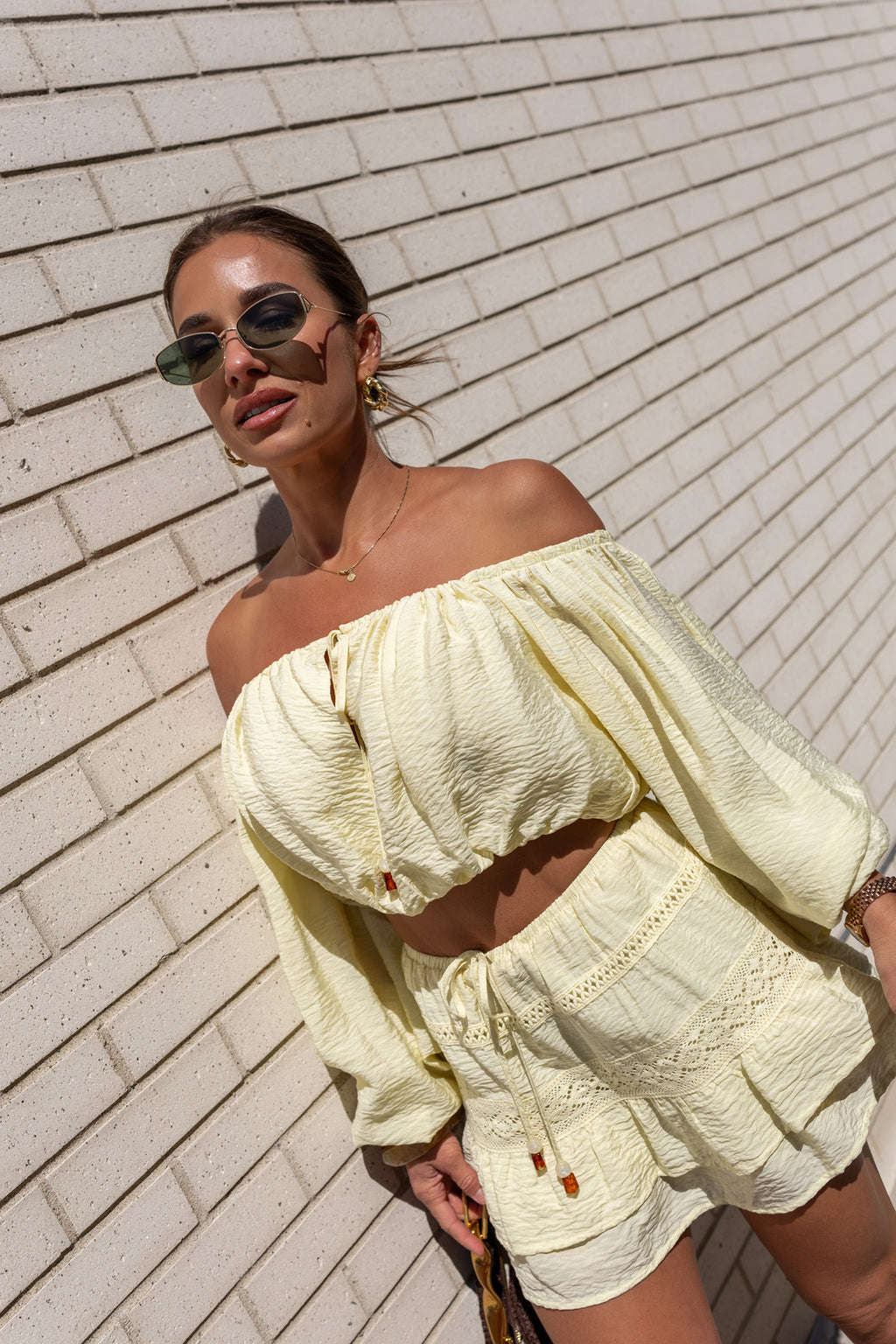 Jessie wearing a light yellow off-shoulder top and skirt against a brick wall.