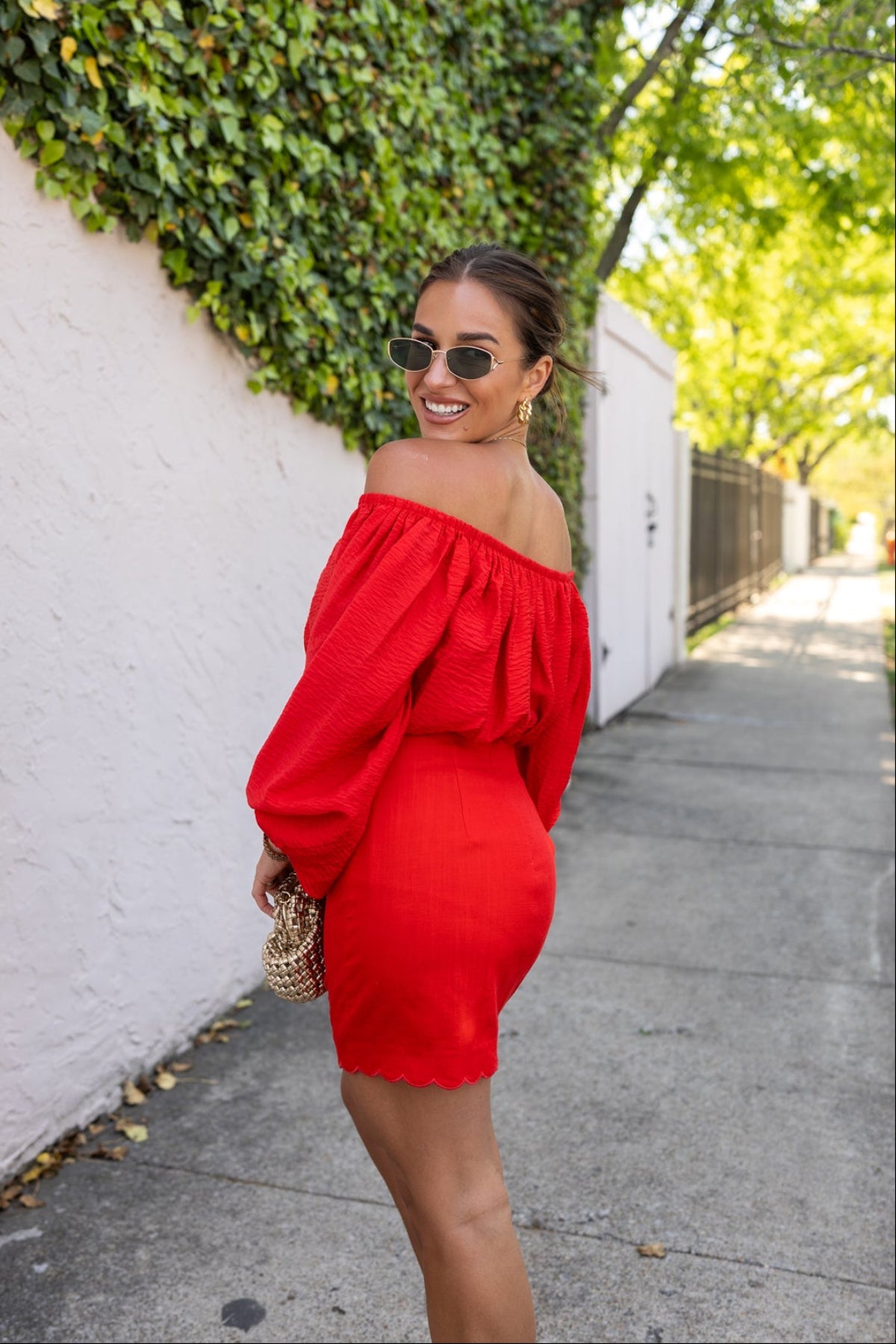 Woman in a red off-shoulder top over a red dress standing against a white wall with greenery.