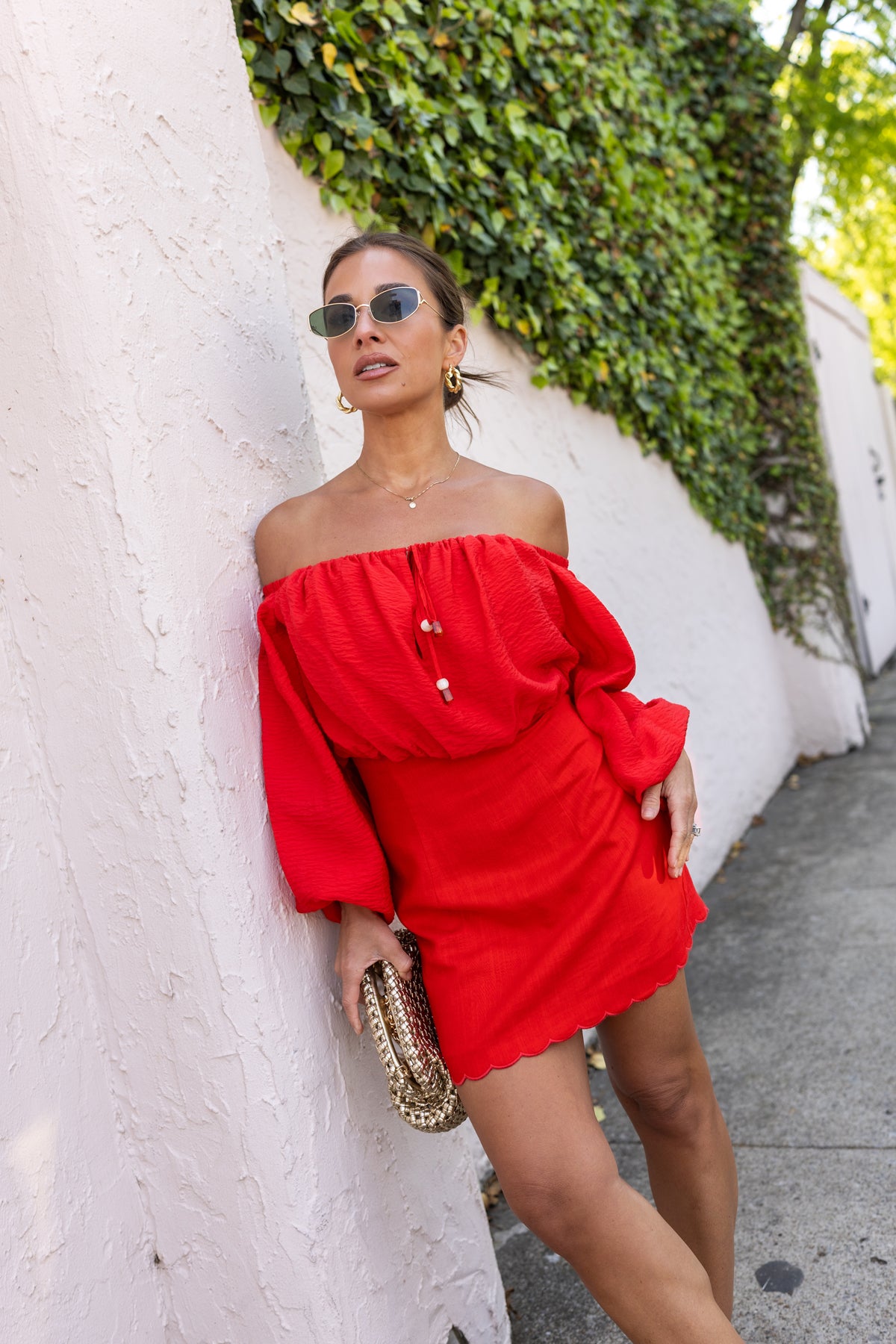 Jessie in a red off-shoulder top over dress standing against a white wall with greenery.