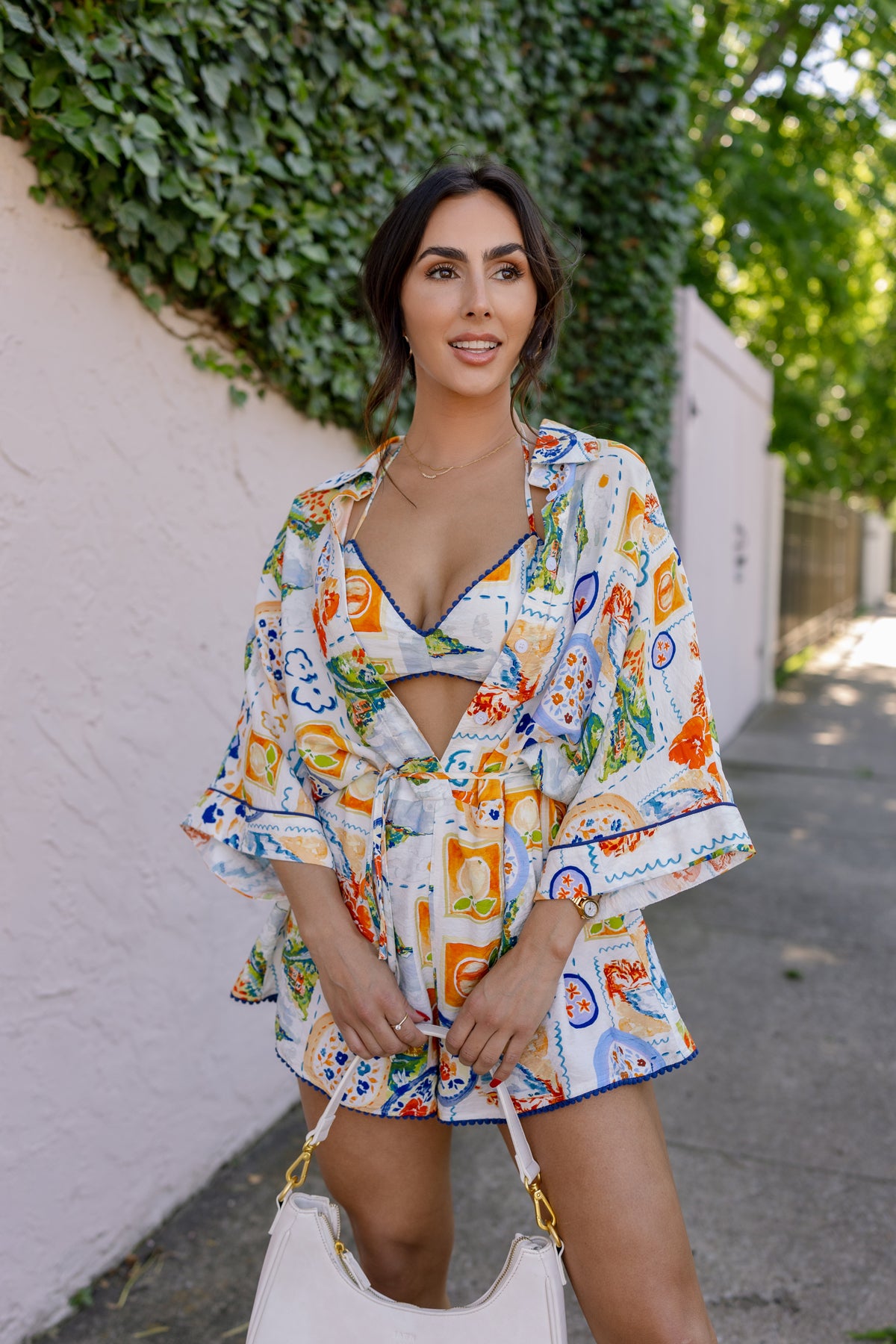 Woman in a colorful floral dress standing on a sidewalk with greenery in the background. Model is wearing the FRANCESCA PRINTED ROMPER WITH BRALETTE IN WHITE.