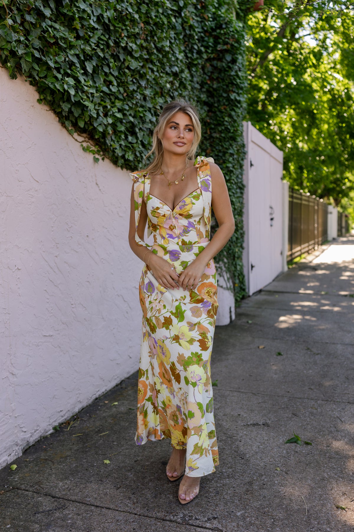 Woman in a floral dress standing against a white wall with greenery. Model is wearing the CATERINA FLORAL MIDI DRESS IN CREAM.