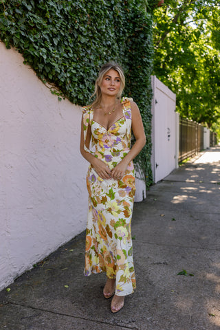 Woman in a floral dress standing against a white wall with greenery. Model is wearing the CATERINA FLORAL MIDI DRESS IN CREAM.