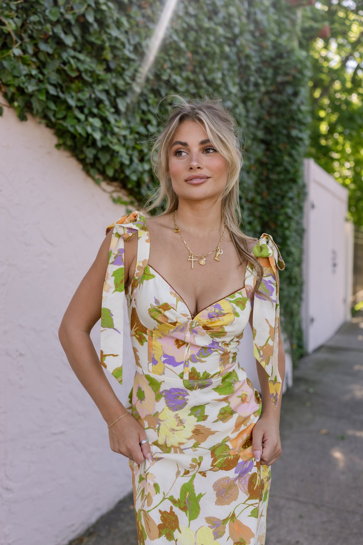 Woman wearing a floral dress standing against a wall with greenery. Model is wearing the CATERINA FLORAL MIDI DRESS IN CREAM.