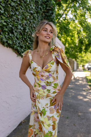 Woman in a floral dress standing outdoors with greenery in the background. Model is wearing the CATERINA FLORAL MIDI DRESS IN CREAM.