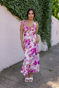 Woman in a floral dress standing against a white wall with greenery. Model is wearing CATERINA FLORAL MIDI DRESS IN WHITE.