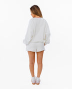 A woman modeling a white crewneck sweatshirt with 'KITTENISH' printed in pink arched letters across the chest, paired with matching white shorts, white socks, and pink sneakers, set against a plain white background.