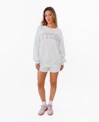 A woman modeling a white crewneck sweatshirt with 'KITTENISH' printed in pink arched letters across the chest, paired with matching white shorts, white socks, and pink sneakers, set against a plain white background.