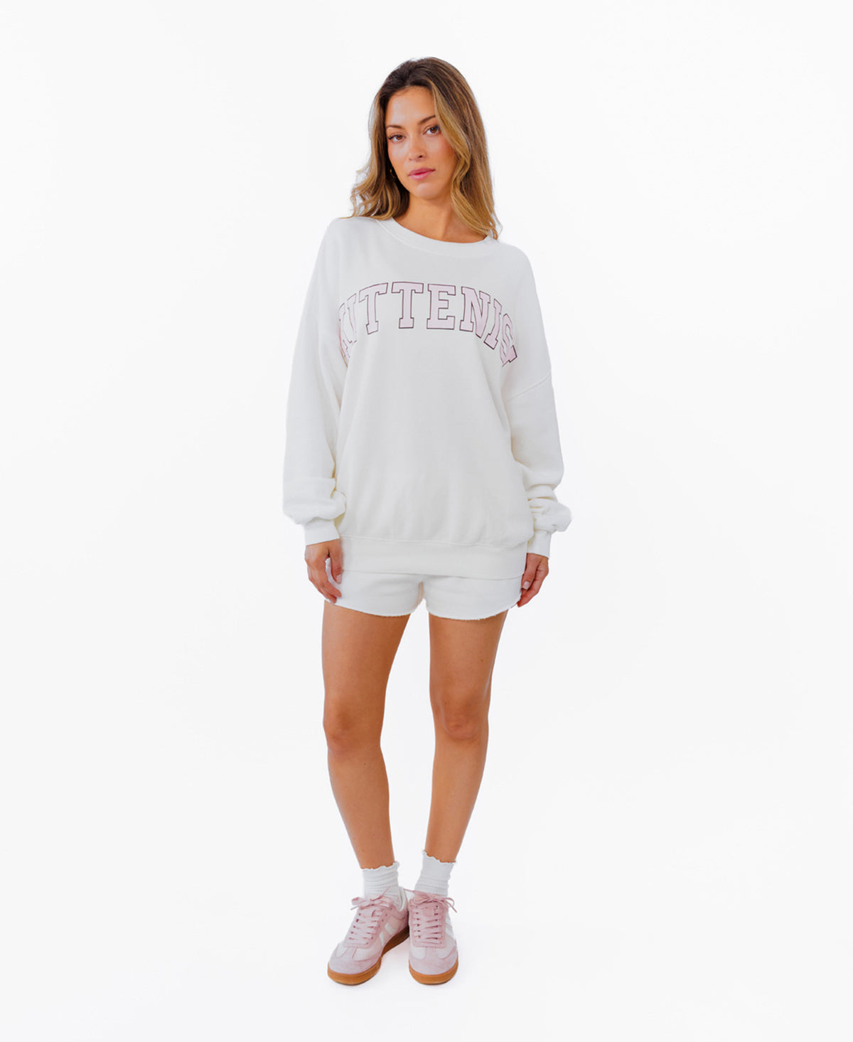 A woman modeling a white crewneck sweatshirt with 'KITTENISH' printed in pink arched letters across the chest, paired with matching white shorts, white socks, and pink sneakers, set against a plain white background.