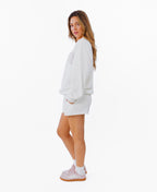 A woman modeling a white crewneck sweatshirt with 'KITTENISH' printed in pink arched letters across the chest, paired with matching white shorts, white socks, and pink sneakers, set against a plain white background.