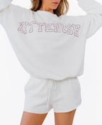A woman modeling a white crewneck sweatshirt with 'KITTENISH' printed in pink arched letters across the chest, paired with matching white shorts, white socks, and pink sneakers, set against a plain white background.