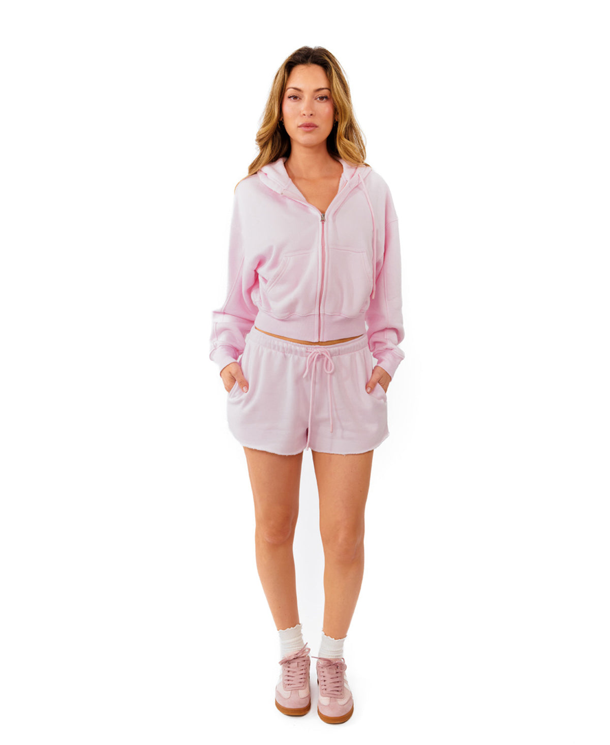 A woman modeling a light pink two-piece lounge set consisting of a cropped zip-up hoodie and matching drawstring shorts, paired with white socks and pink sneakers, against a solid white background.