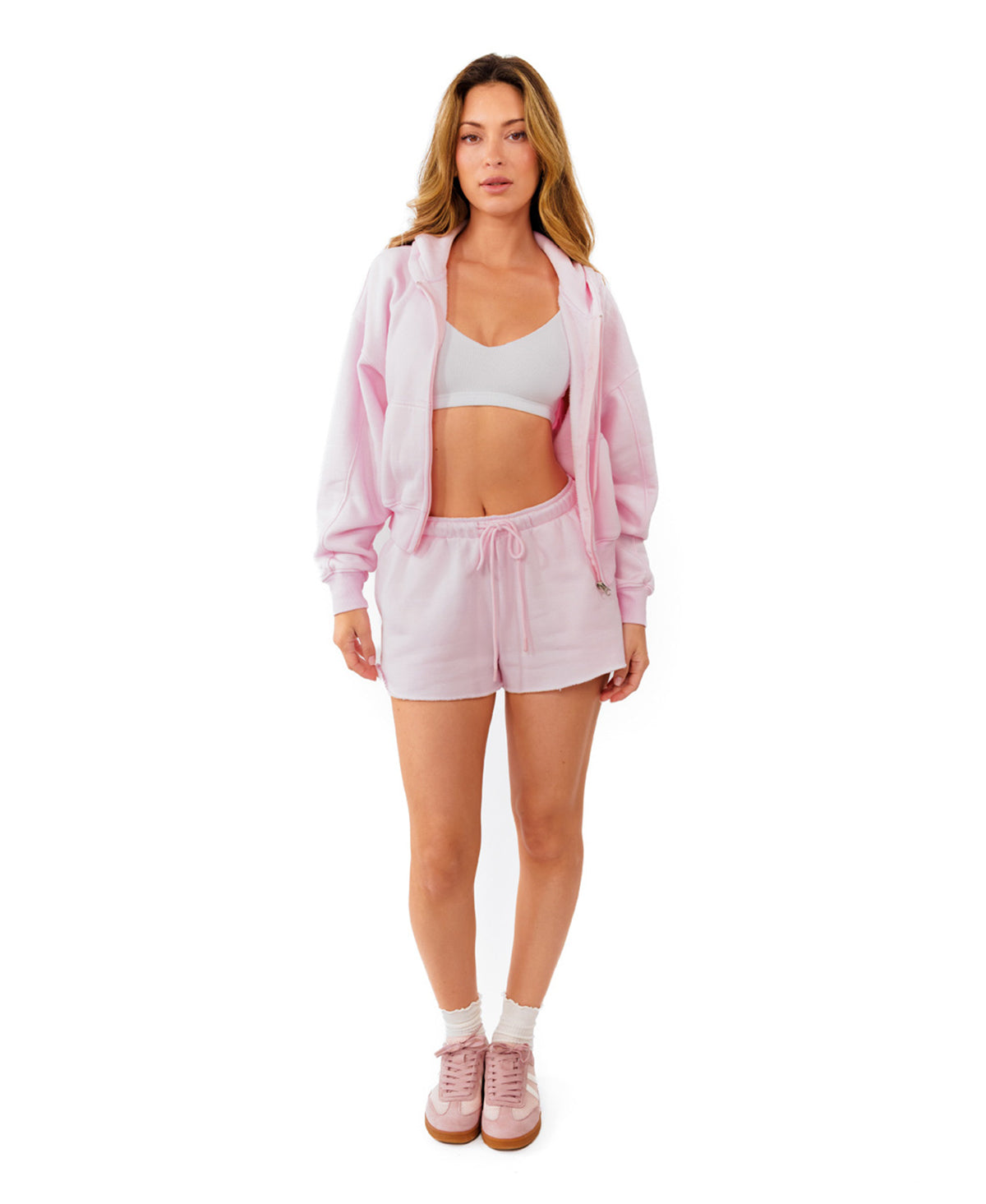 A woman modeling a light pink two-piece lounge set consisting of a cropped zip-up hoodie and matching drawstring shorts, paired with white socks and pink sneakers, against a solid white background.