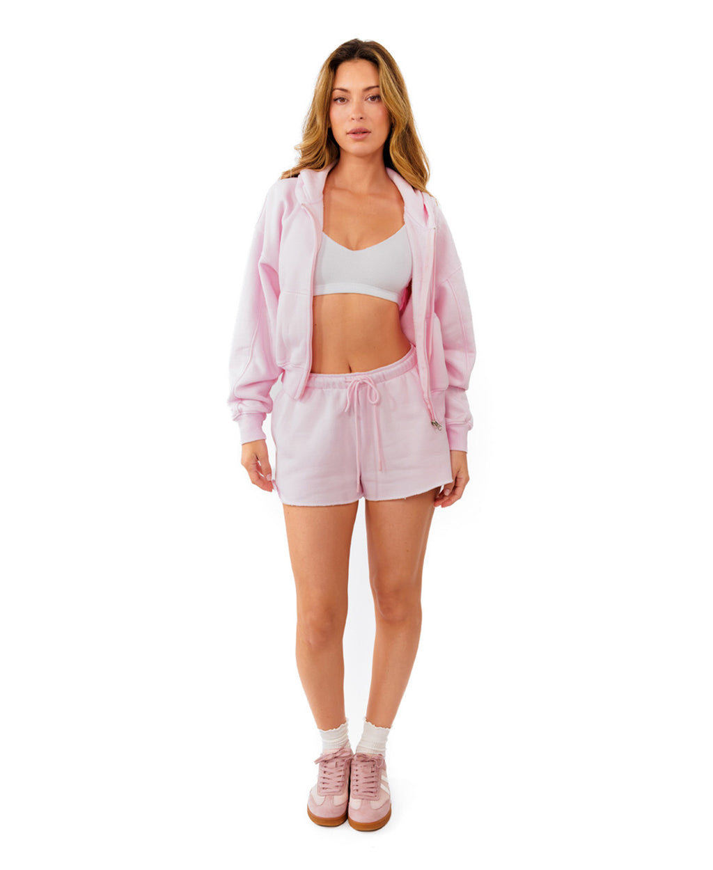 A woman modeling a light pink two-piece lounge set consisting of a cropped zip-up hoodie and matching drawstring shorts, paired with white socks and pink sneakers, against a solid white background.