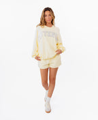 A woman modeling a butter yellow lounge set consisting of a sweatshirt with 'KITTENISH' printed in white across the chest and matching shorts, styled with white socks and white sneakers against a plain white background.