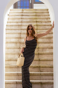 Jessie wearing a black and white horizontally striped sleeveless maxi dress stands on a stone staircase, holding a beige woven tote bag.