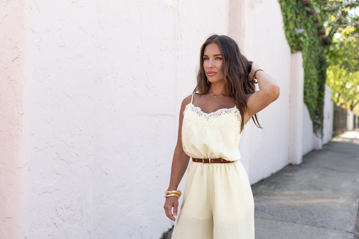 Woman in a light yellow jumpsuit standing against a white wall with greenery.
