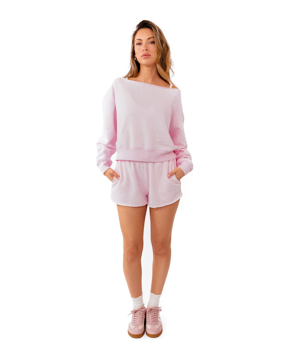 A woman modeling a light pink two-piece loungewear set consisting of an off-the-shoulder long-sleeve sweatshirt and matching shorts, paired with white socks and pink sneakers, against a solid white background.