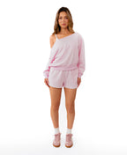 A woman modeling a light pink two-piece loungewear set consisting of an off-the-shoulder long-sleeve sweatshirt and matching shorts, paired with white socks and pink sneakers, against a solid white background.