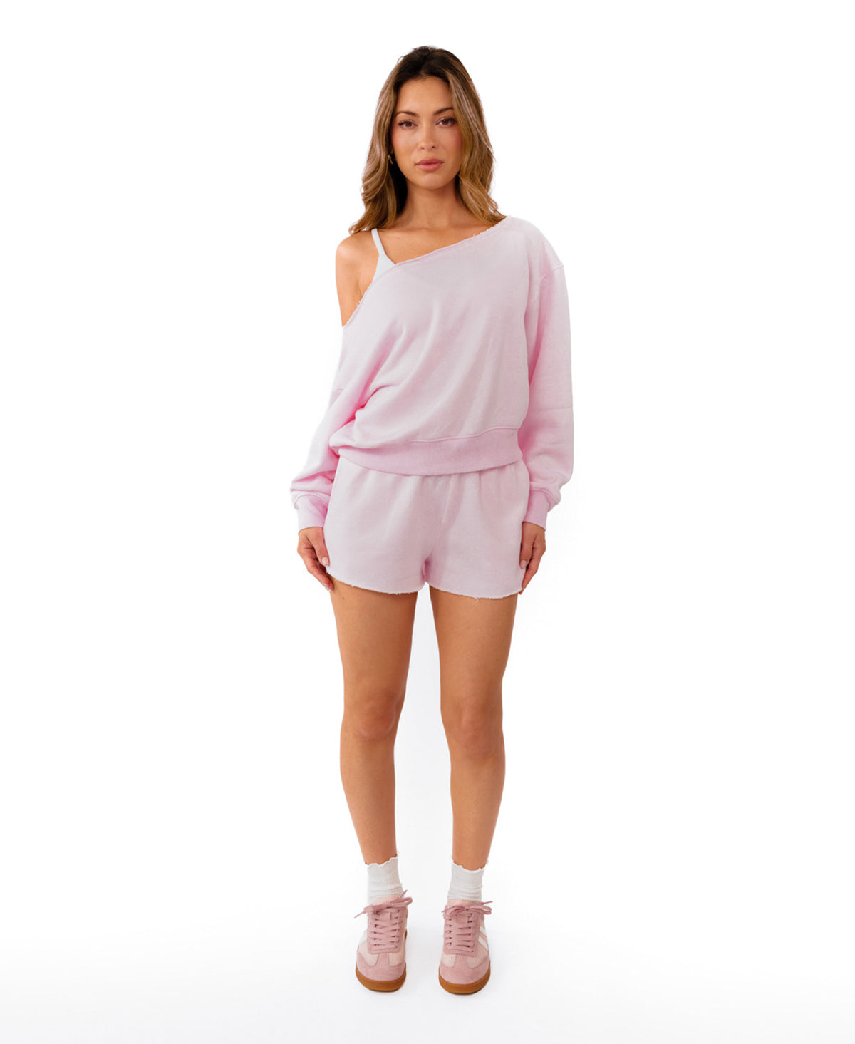 A woman modeling a light pink two-piece loungewear set consisting of an off-the-shoulder long-sleeve sweatshirt and matching shorts, paired with white socks and pink sneakers, against a solid white background.