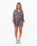 A woman modeling a matching two-piece lounge set consisting of a zip-up hooded sweatshirt and drawstring shorts in a heathered  gray color, styled with white socks and sneakers against a plain white background.
