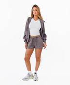 A woman modeling a matching gray lounge set consisting of a zip-up hoodie and drawstring shorts, styled with a white ribbed crop top, white socks, and gray sneakers against a plain white background.