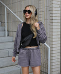 A woman stands on outdoor concrete stairs wearing a matching gray lounge set consisting of a zip-up hoodie and shorts, a black crop top, a black crossbody bag, and black sunglasses.
