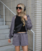 A woman stands on outdoor concrete stairs wearing a matching gray lounge set consisting of a zip-up hoodie and shorts, a black crop top, a black crossbody bag, and black sunglasses.