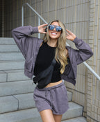 A woman posing on outdoor concrete stairs wearing a matching heather purple lounge set consisting of a cropped zip-up hoodie and drawstring shorts, a black tank top, and a black crossbody bag.