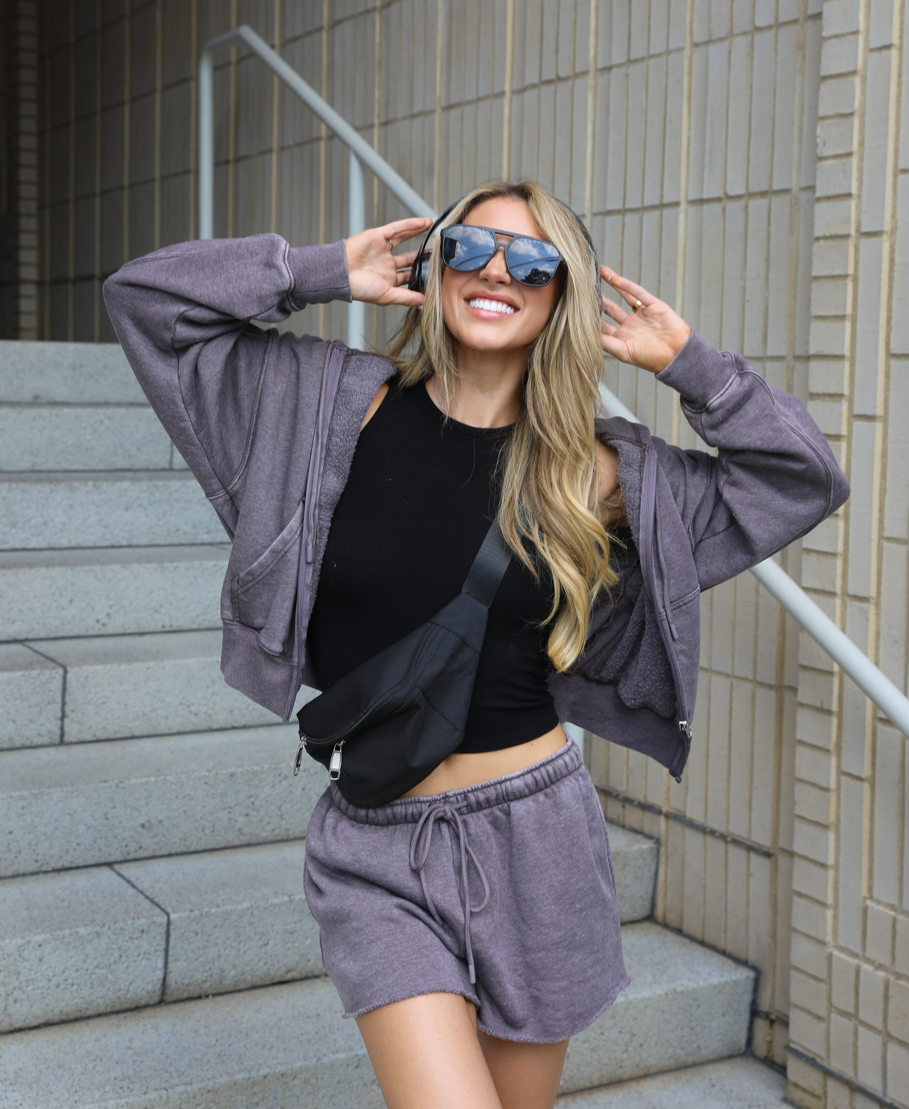 A woman posing on outdoor concrete stairs wearing a matching heather purple lounge set consisting of a cropped zip-up hoodie and drawstring shorts, a black tank top, and a black crossbody bag.