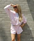 A woman wearing a light pink zip-up hoodie and matching shorts over a white t-shirt, posing against a gray brick wall.