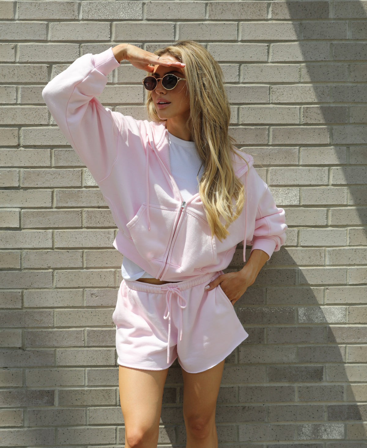 A woman wearing a light pink zip-up hoodie and matching shorts over a white t-shirt, posing against a gray brick wall.