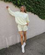 A woman posing outdoors against a stucco wall with ivy, wearing a butter yellow 'KITTENISH' sweatshirt and matching shorts, white sneakers, and a black patterned headband.