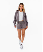 A woman modeling a matching gray lounge set consisting of a cropped zip-up hoodie and drawstring shorts, styled with a white ribbed crop top and white sneakers against a plain white background.