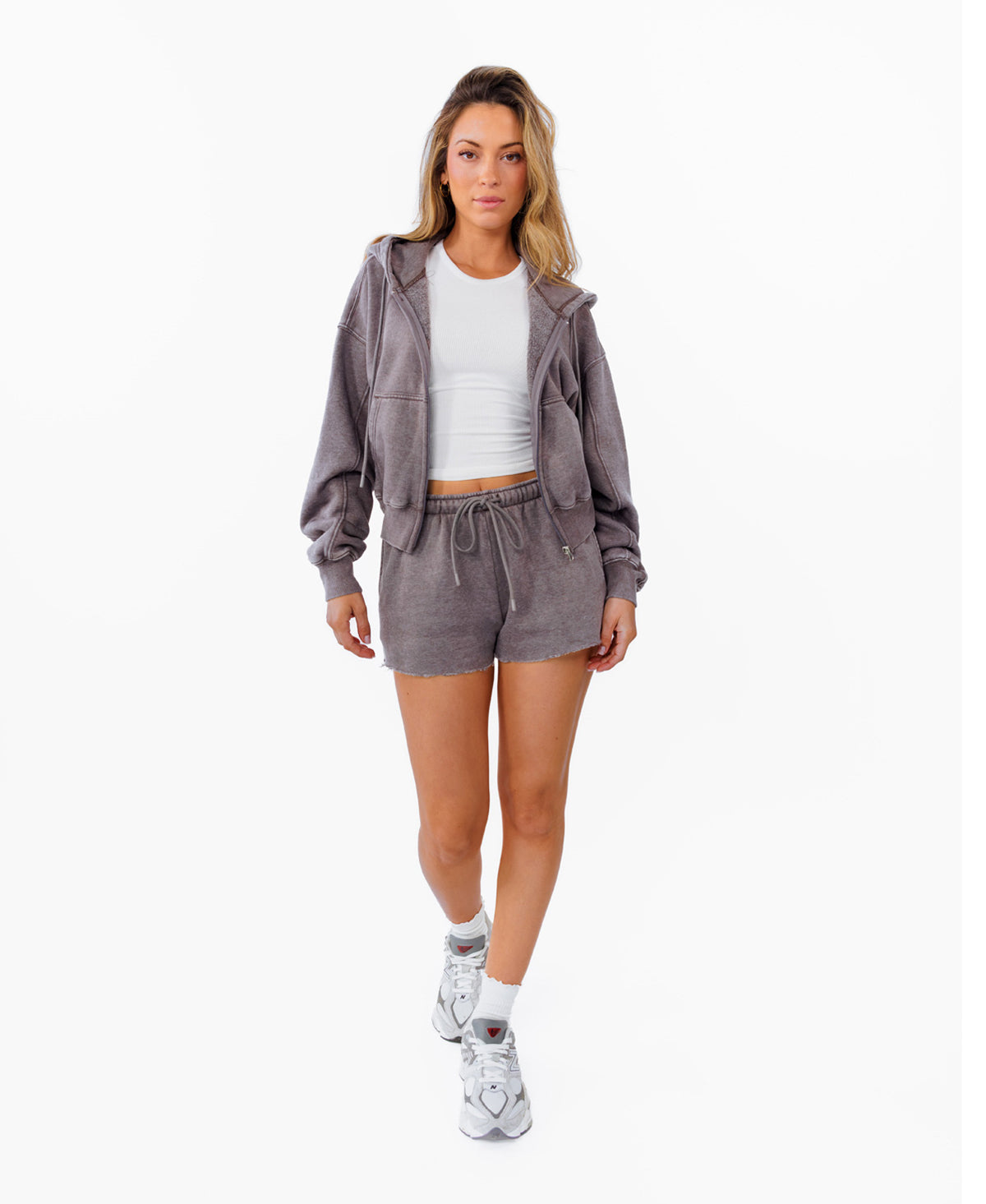 A woman modeling a matching gray lounge set consisting of a cropped zip-up hoodie and drawstring shorts, styled with a white ribbed crop top and white sneakers against a plain white background.