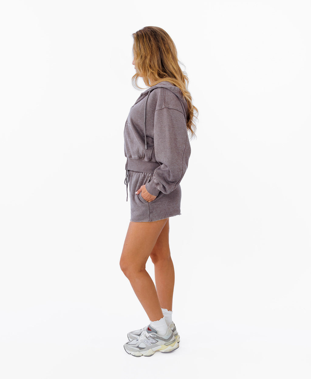 A woman stands in profile wearing a matching heathered  gray hoodie and shorts set, white socks, and chunky white sneakers against a plain white studio background.