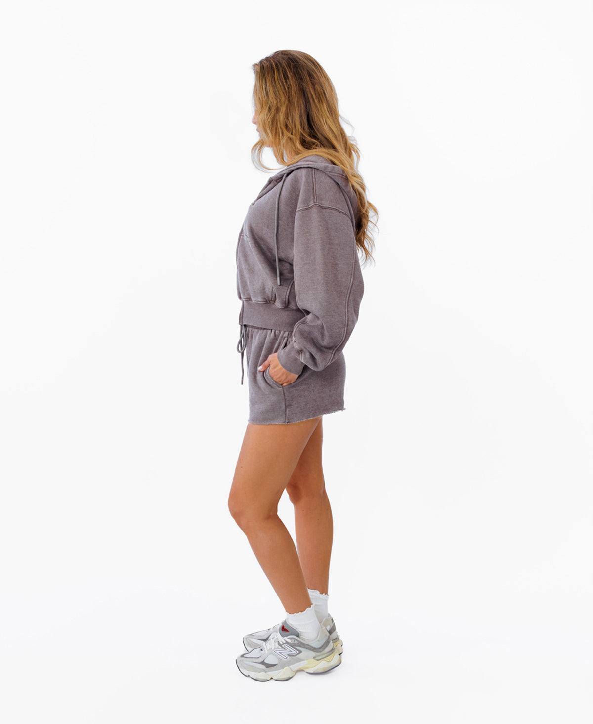 A woman stands in profile wearing a matching heathered  gray hoodie and shorts set, white socks, and chunky white sneakers against a plain white studio background.