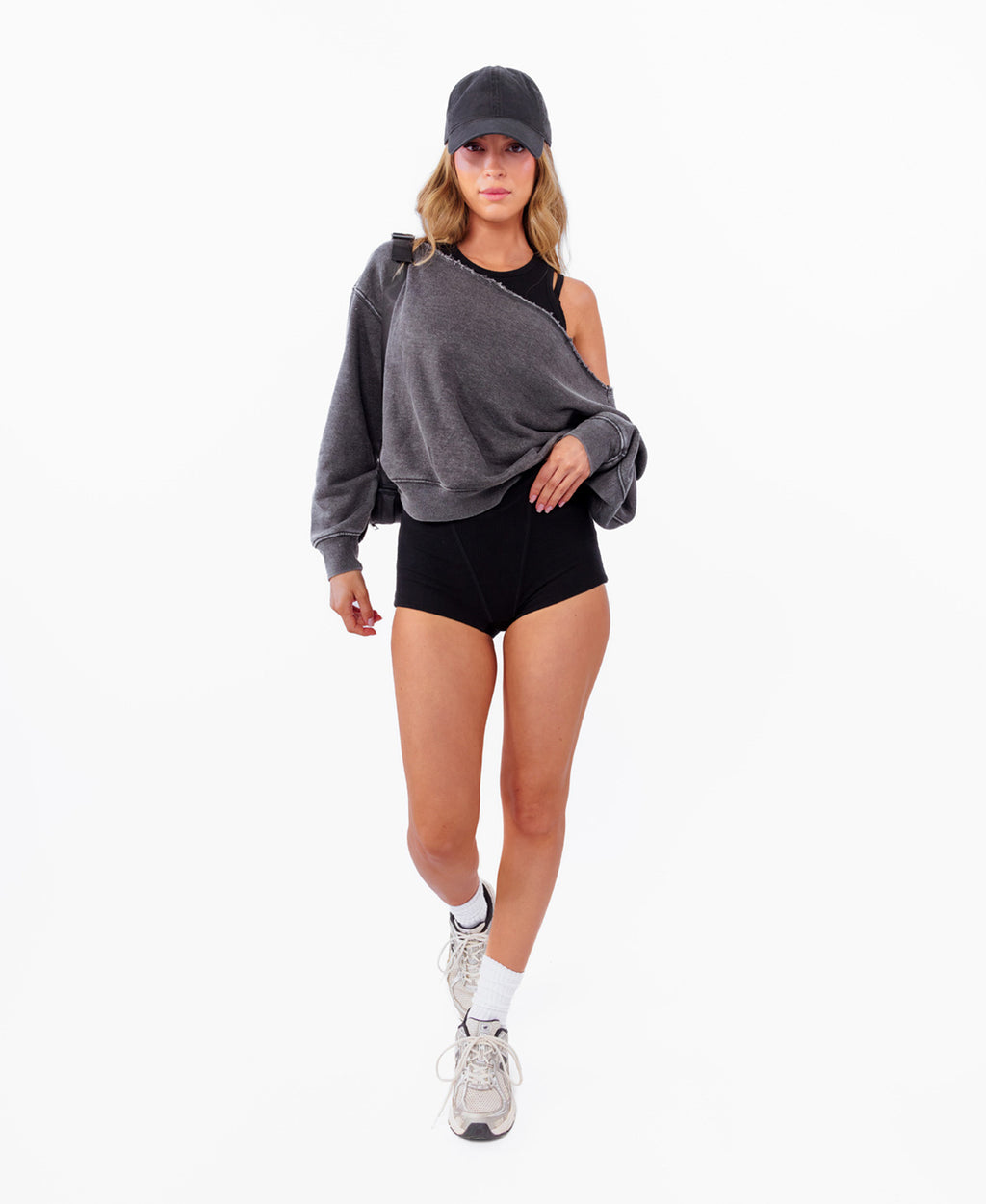 A model wearing a dark gray oversized off shoulder sweatshirt, black athletic shorts, a black cap, white socks, and gray sneakers, standing against a plain white background.